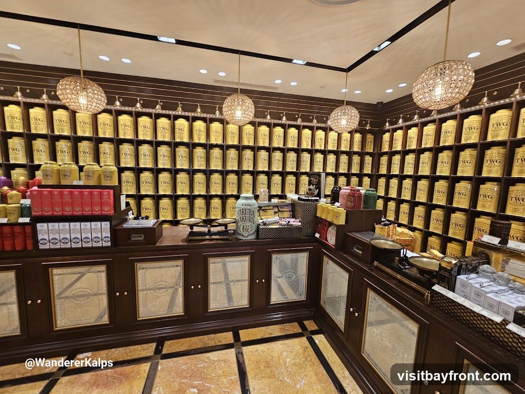A well-lit interior of a tea shop, with walls lined with shelves filled with numerous yellow tins of tea. A wooden counter runs along the front, displaying various tea-related items, including scales, boxes, and decorative tins. The overall ambiance is elegant and sophisticated.