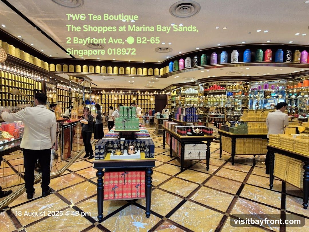 The interior of a TWG Tea Boutique, showcasing rows of tea canisters on shelves and display tables laden with tea boxes. Several staff members in uniform are visible assisting customers. The store features ornate decor and a marble-tiled floor.