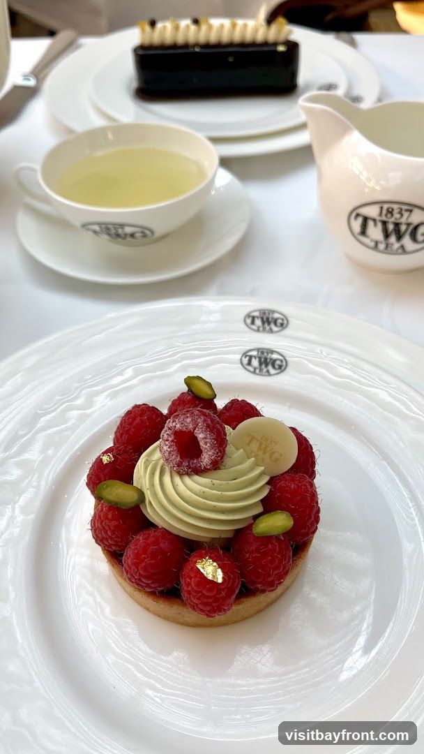A close-up of a raspberry tart with pistachio cream and gold leaf, served on a white plate with the TWG Tea logo. A cup of tea and a small pitcher, also branded with TWG Tea, are visible in the background, along with a dark chocolate dessert.