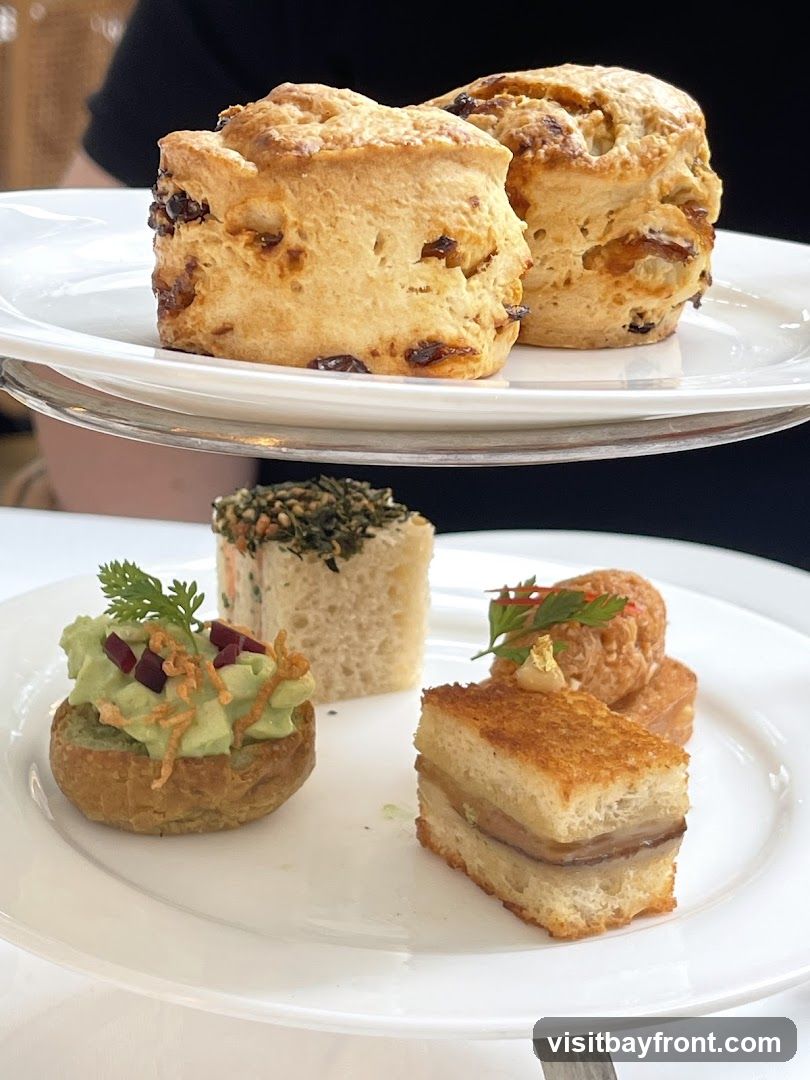 A tiered stand holds an assortment of pastries and scones. The top tier features two golden-brown scones studded with raisins. The lower tier displays four delicate finger foods: a small pastry shell filled with avocado and topped with microgreens and crispy onions, a square sandwich with a green topping, a small round item with a garnish, and a layered sandwich with toasted bread.