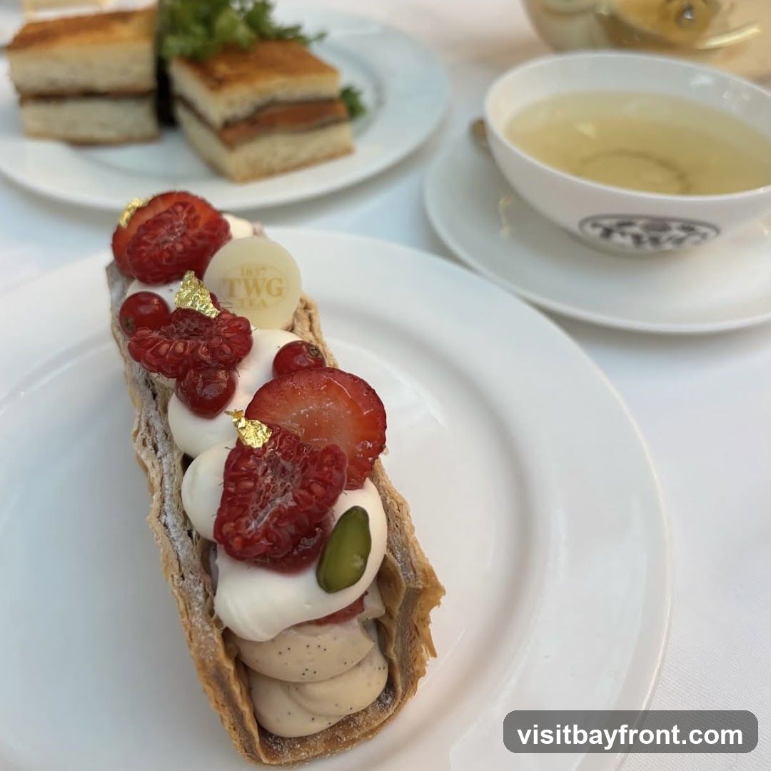 A close-up of a long, thin pastry filled with cream and topped with fresh berries, pistachios, and edible gold leaf. In the background, a cup of tea and two small sandwiches are visible on white plates. The pastry has a TWG Tea logo on a white chocolate disc.