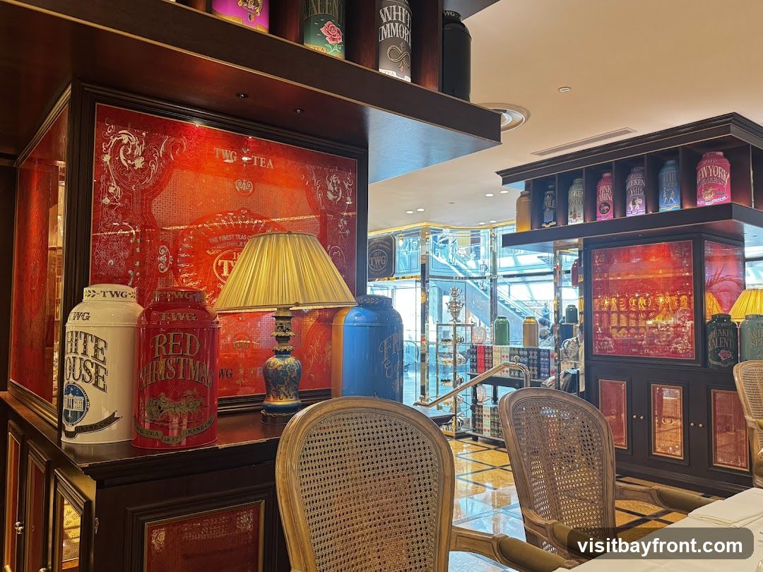 The image shows an interior view of a tea shop. Prominently displayed are large decorative tins labeled with tea names such as 'TWG WHITE HOUSE' and 'TWG RED CHRISTMAS'. A lamp with a golden shade sits on a shelf. In the background, more tins are visible on shelves, along with a glimpse of a staircase and other patrons. Wicker chairs are set at a table covered with a white tablecloth. The decor features rich red accents and ornate details.