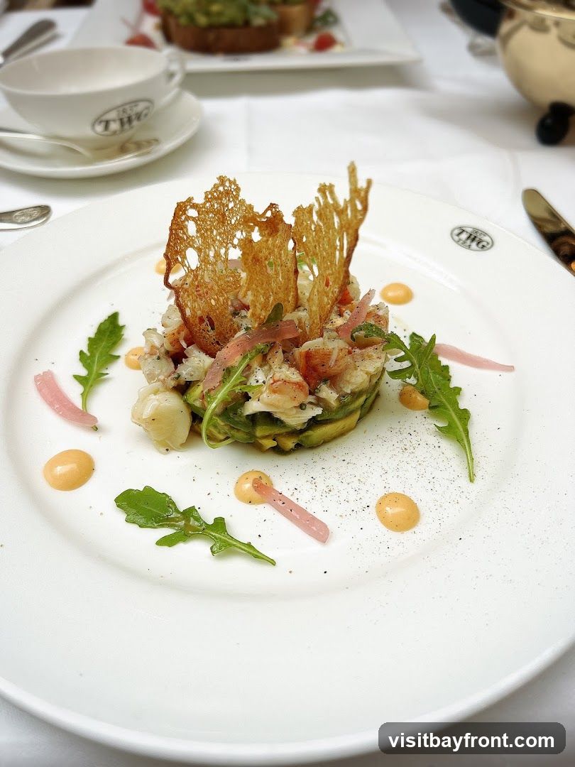 A close-up shot of a seafood and avocado salad served on a white plate. The salad is topped with a crispy lattice cracker and garnished with arugula leaves, pickled red onions, and dollops of sauce. A teacup with a logo is visible in the background.