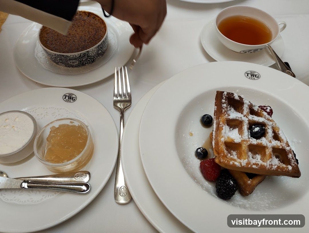 A close-up view of a table setting with various desserts and a cup of tea. In the foreground, a plate holds a waffle topped with powdered sugar and fresh berries, accompanied by a small dish of jam and a dollop of cream. To the left, a crème brûlée is presented in a decorative bowl. A cup of tea sits on a saucer in the upper right corner. The TWG Tea logo is visible on the plates and cup.