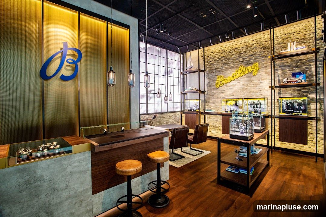 The interior of a Breitling watch store features a modern and luxurious design. A large blue Breitling logo is displayed on a gold textured wall. The store has a wooden counter with display cases showcasing watches. There are also seating areas with leather chairs and stools. Shelving units display watches, models, and promotional materials. The walls are decorated with exposed brick and neon signage that reads "Breitling".