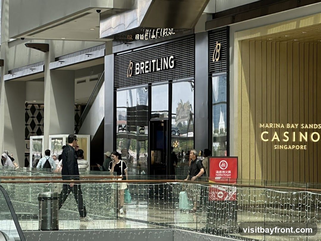 A view of a Breitling store with its name prominently displayed above the entrance. To the right, the entrance to the Marina Bay Sands Casino in Singapore is visible. People are walking in the foreground on a walkway with a railing.
