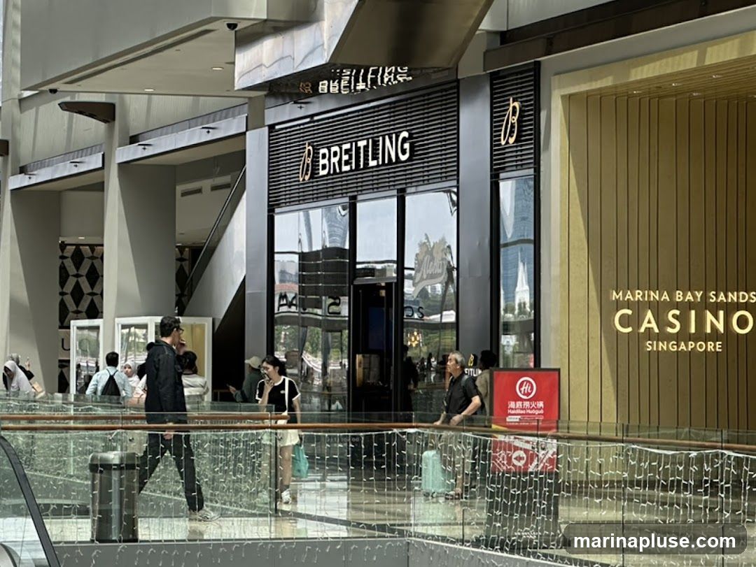 A view of a Breitling store with its name prominently displayed above the entrance. To the right, the entrance to the Marina Bay Sands Casino in Singapore is visible. People are walking in the foreground on a walkway with a railing.