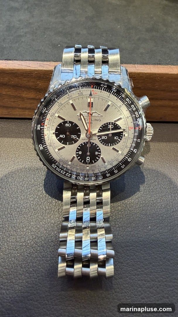 A close-up shot of a Breitling Navitimer watch with a silver dial and a stainless steel bracelet. The watch features multiple subdials and a slide rule bezel. It is displayed on a dark textured surface with a wooden edge in the background.