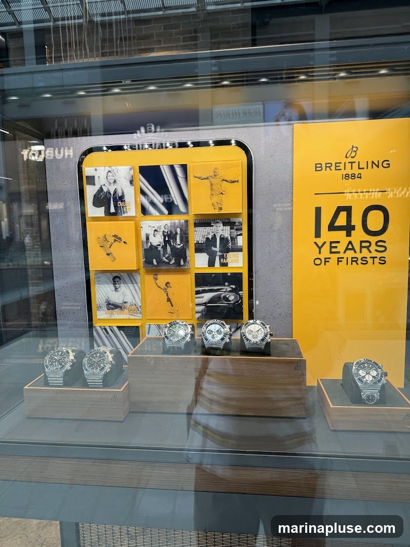 A display window showcasing Breitling watches. A large yellow sign reads "BREITLING 1884 140 YEARS OF FIRSTS". A grid of nine yellow squares features images of people and watches, including Chloe Kim and Erling Haaland. Several Breitling watches are displayed on wooden stands in the window.