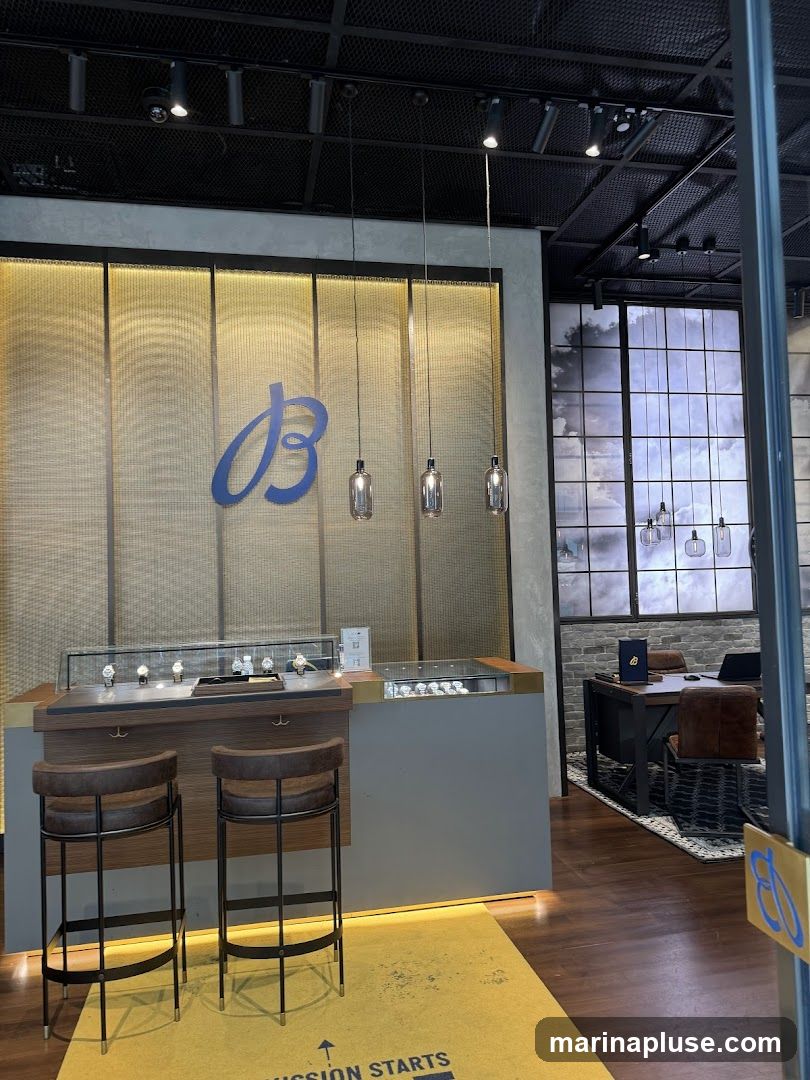 The interior of a Breitling watch store features a display counter with watches, two bar stools, and a large blue Breitling logo on a textured wall. To the right, a desk area with chairs is visible, along with a window displaying a cloudy sky. A yellow mat with text is on the floor.