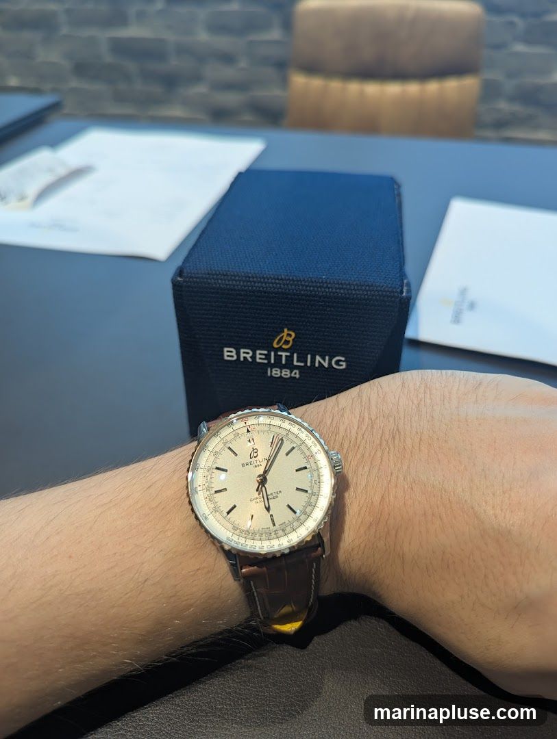 A close-up shot of a Breitling watch on a person's wrist, with a Breitling watch box in the background. The watch has a silver dial and a brown leather strap. The box is dark blue with the Breitling logo and "1884" printed on it. There are also some papers on the table.