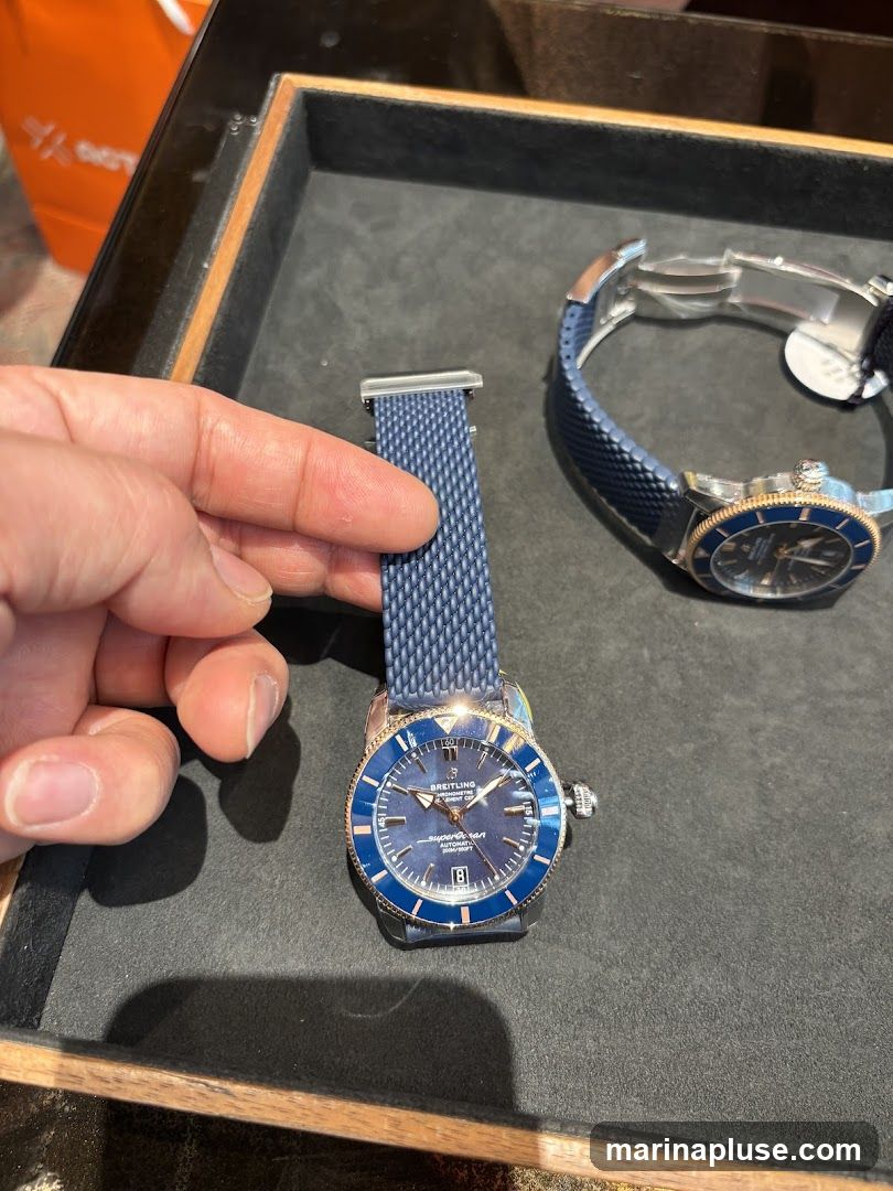 A hand is holding a blue mesh watch strap next to a Breitling Superocean automatic watch with a blue dial and rose gold accents. Another watch with a blue strap is visible in the background. The watches are displayed on a dark gray surface within a wooden tray. An orange shopping bag is partially visible in the upper left corner.