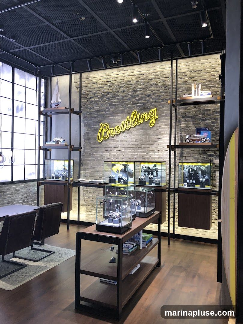 The interior of a Breitling watch store features a brick wall with a neon yellow "Breitling" sign. Display cases showcase watches, and shelves hold books and decorative items like a model sailboat and a vintage car. A table with chairs is visible in the foreground.