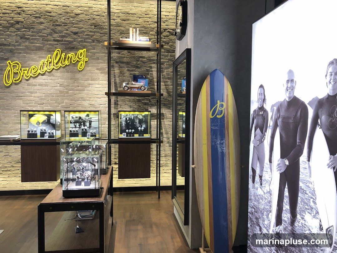 The interior of a Breitling store features a neon sign with the brand name, display cases with watches, shelves with model cars and books, and a surfboard. A large black and white photograph of surfers is displayed on the right side of the store.