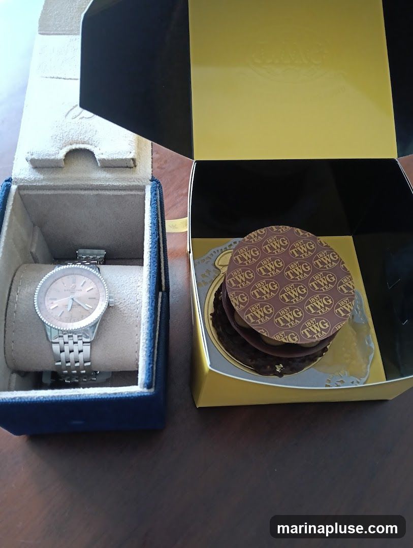 A silver watch with a rose gold face is displayed in a blue velvet box. Next to it, a yellow box contains a chocolate cake with a decorative paper lid featuring the TWG Tea logo. The cake appears to be layered and has gold leaf accents.