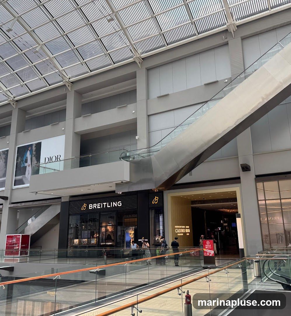 A view of a Breitling watch store and a casino entrance within a modern shopping mall. The Breitling store has a black facade with the brand name and logo prominently displayed. Inside, watches are visible in display cases. Adjacent to the Breitling store is the entrance to a casino, marked with signage in multiple languages. An escalator is visible in the background, along with a large DIOR advertisement on an upper level. The mall features a glass ceiling and multiple levels with glass railings.