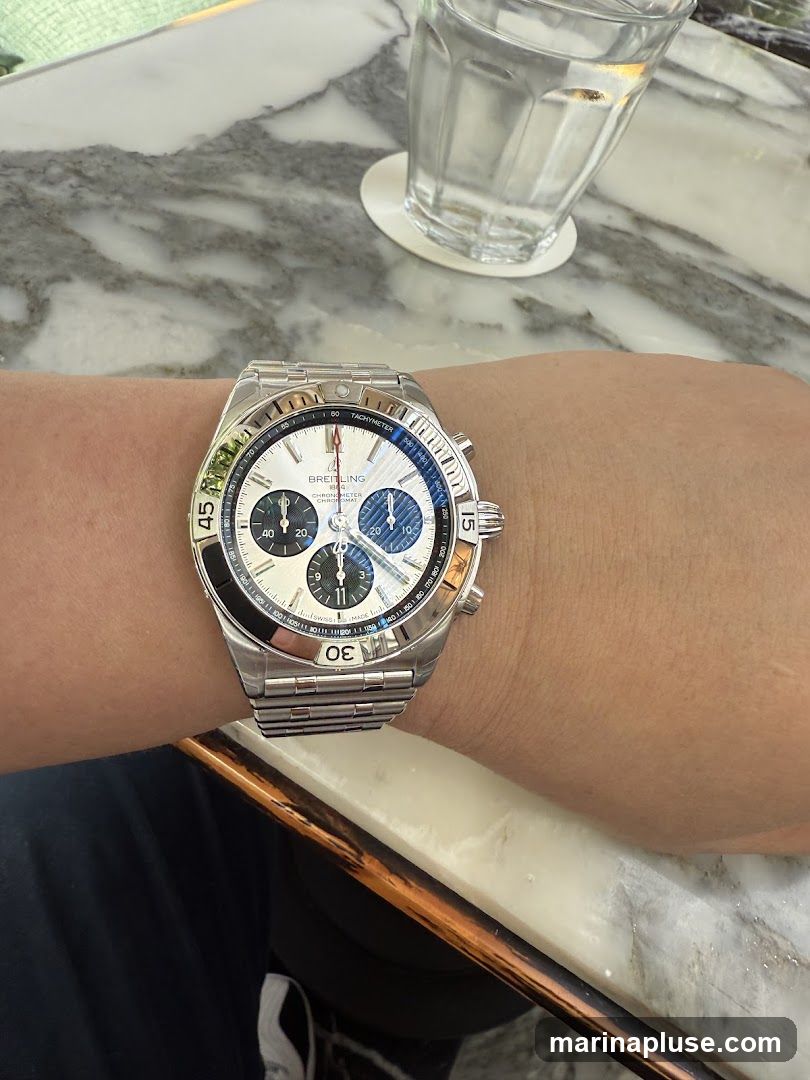 A close-up shot of a person's wrist wearing a Breitling Chronomat watch. The watch has a silver metal band and a white face with blue and black sub-dials. The bezel is black and silver with blue markings. In the background, a glass of water sits on a coaster on a marble-patterned table.