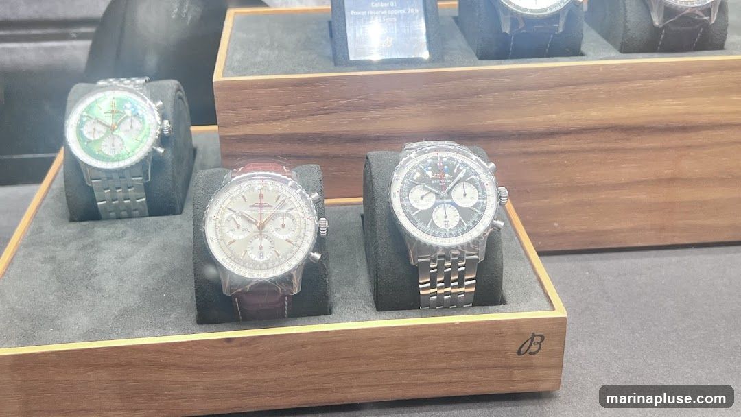 A display of several luxury watches, each on a dark grey velvet stand within a wooden case. The watches feature various dial colors, including green and silver, and metal or leather straps. One watch has a prominent green dial with three sub-dials and a metal bracelet. Another has a silver dial with a brown leather strap. A third watch, partially visible, has a dark dial and a metal bracelet. A small plaque above the watches displays text, including "Caliber 01" and "Power reserve approx. 70 h". The Breitling logo is visible on the wooden case.