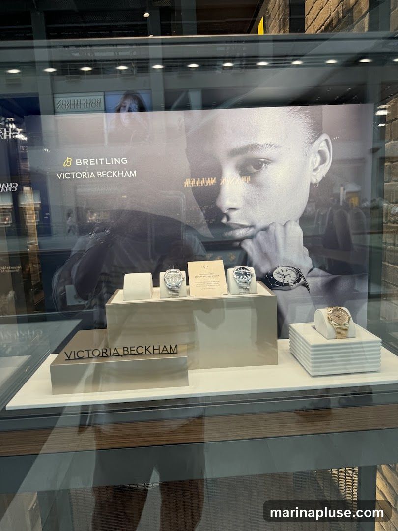 A display case featuring Breitling watches, with a large black and white advertisement of a woman's face and the logos for Breitling and Victoria Beckham. Several watches are showcased on pedestals, including a gold-toned watch on the right. The display is behind glass, with reflections visible.