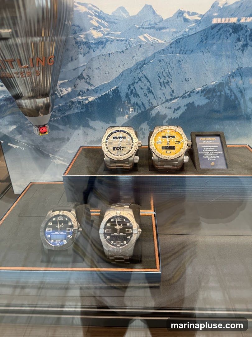 A display case featuring several Breitling watches against a backdrop of snowy mountains. In the foreground, two watches are shown on the lower level, one with a black strap and blue digital display, and another with a metallic band and black analog face. On the upper level, two more watches are displayed, one silver and one yellow, with digital and analog faces. A small placard next to the watches reads "PROFESSIONAL EMERGENCY" and describes a Breitling Caliber 76. A silver, cylindrical object with "BREITLING ORBITER 3" visible on it is positioned in the upper left corner.