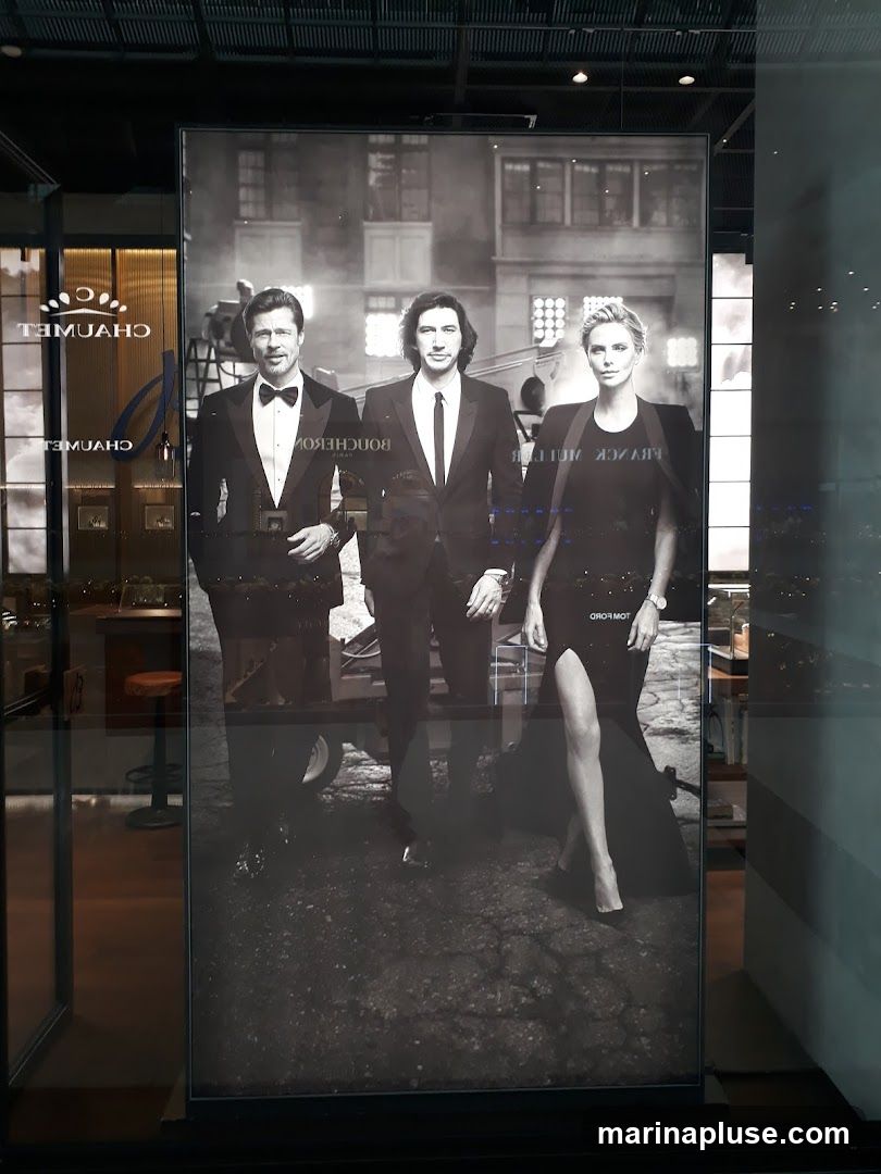 A black and white advertisement featuring actors Brad Pitt, Adam Driver, and Charlize Theron. They are dressed in formal attire. The advertisement appears to be for Boucheron, as the brand name is visible on the image. The advertisement is displayed in a window, with reflections of the interior and exterior visible.