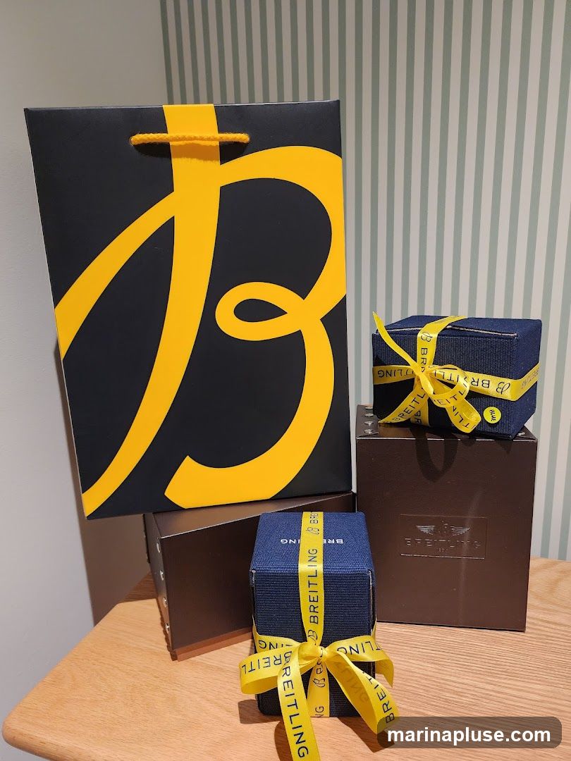 A large black gift bag with a large yellow stylized 'B' logo is placed on top of a dark brown box. To the right, two smaller dark blue boxes are tied with yellow ribbon featuring the Breitling logo. These boxes are also placed on top of dark brown boxes, one of which has the Breitling logo embossed on it. The items are arranged on a wooden surface against a striped wallpaper background.