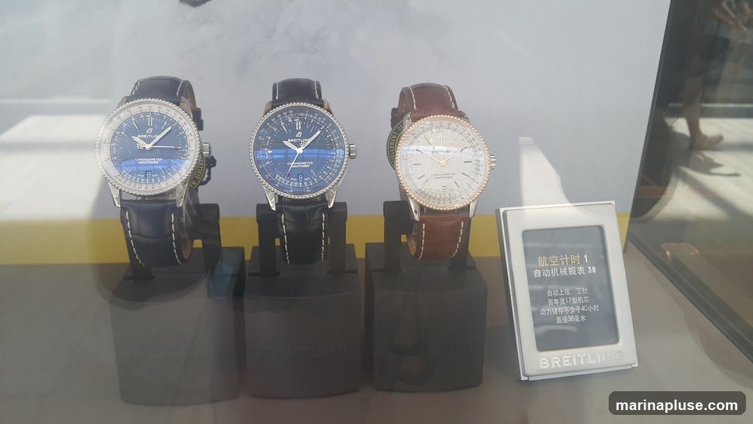 Three Breitling watches are displayed on stands. Two watches have blue dials and black leather straps, while the third has a white dial and a brown leather strap. A small digital display with Chinese text and the Breitling logo is in the foreground.