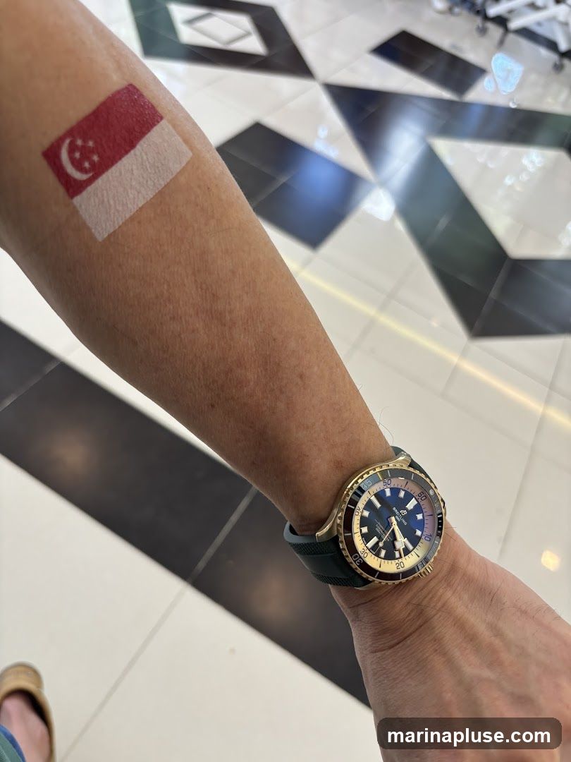 A close-up shot of a person's arm displaying a temporary tattoo of the Singapore flag on the upper arm. A gold-toned watch with a dark green strap is worn on the wrist. The background shows a black and white geometric tiled floor.