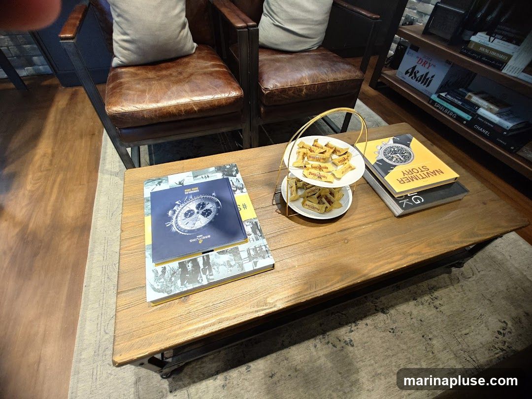 A wooden coffee table holds a two-tiered stand with individually wrapped candies, two books, and a magazine. The books appear to be about watches, with one titled "Navitimer Story." In the background, two leather armchairs with light-colored cushions are visible, along with a bookshelf filled with various books and decorative items.