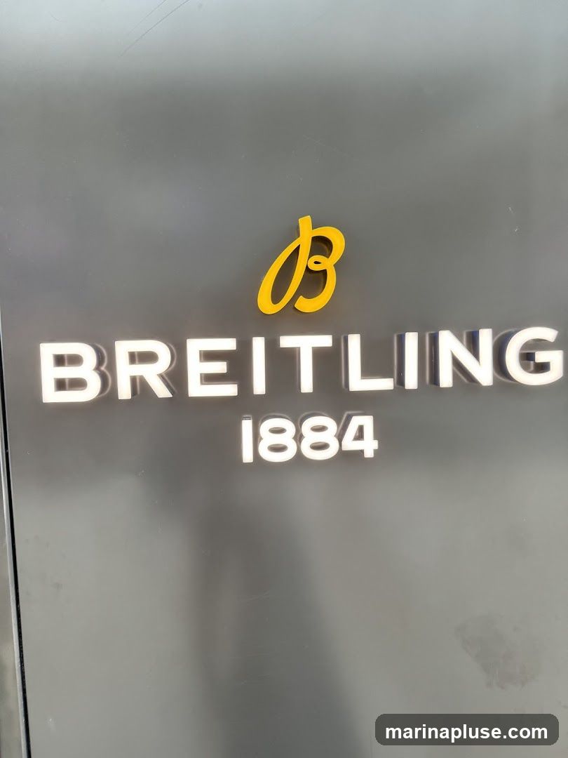 A close-up shot of the Breitling logo and name displayed on a dark gray surface. The logo, a stylized yellow 'B', is positioned above the white, illuminated text 'BREITLING' and the number '1884'.