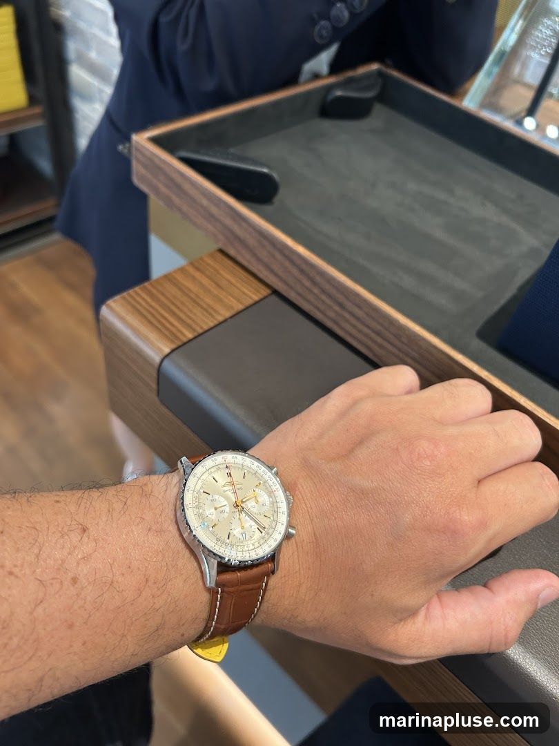 A person's wrist wearing a silver watch with a brown leather strap. The watch face is cream-colored with gold accents and multiple subdials. The background shows a display tray and a person in a dark suit.