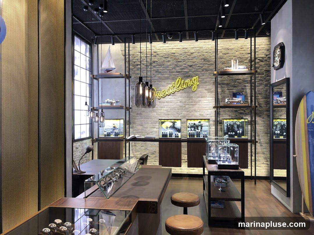 The interior of a Breitling watch store features a brick wall with a neon sign that says "Breitling." Display cases showcase watches, and shelves hold various items, including model airplanes and cars. A surfboard is also visible near a mirror.