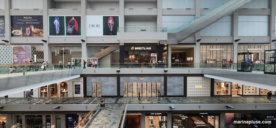 The image shows the interior of a modern shopping mall with multiple levels. Several high-end retail stores are visible, including Breitling, Mikimoto, Dior, and Gucci. Large advertisements for brands like Wakuda and Dior are displayed on the upper levels. Escalators and walkways connect the different floors, and shoppers can be seen browsing the stores. The architecture is sleek and contemporary, with glass railings and polished floors.