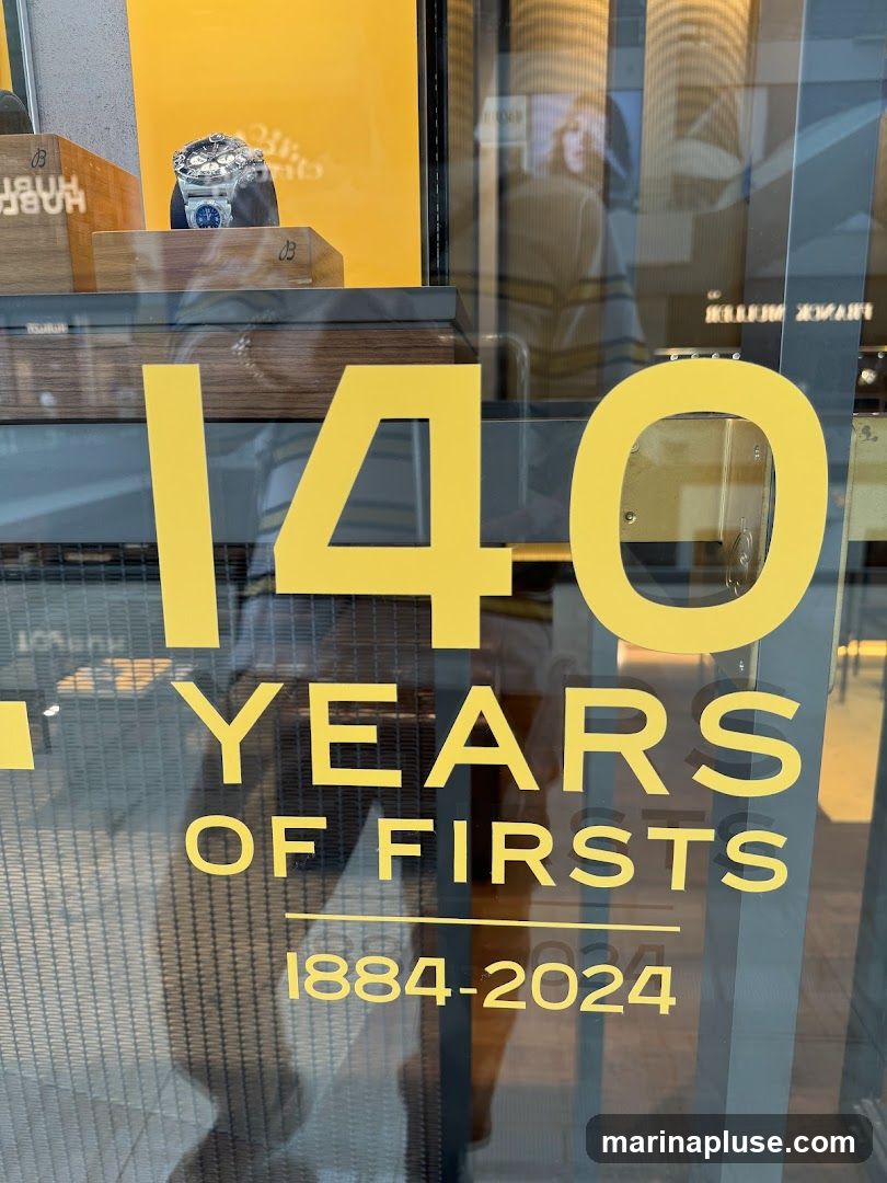 A close-up view of a watch store's window display, featuring large yellow text that reads "140 YEARS OF FIRSTS 1884-2024". A luxury watch is displayed on a wooden stand inside the window. Reflections on the glass show a person wearing a striped shirt.