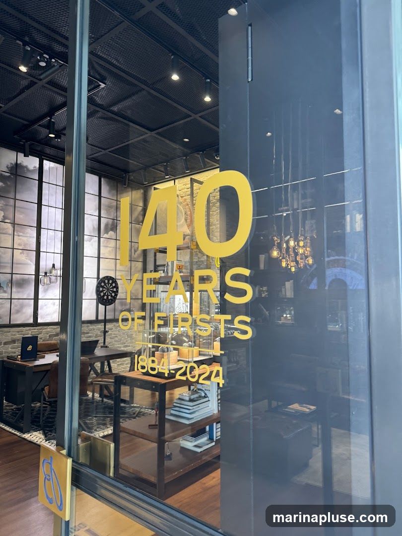 A view through a glass door into a store. Large yellow lettering on the glass reads "140 YEARS OF FIRSTS 1884-2024". Inside, the store appears to be a retail space with shelves displaying books and merchandise. A desk with a laptop and a dartboard are also visible. The ceiling has exposed beams and track lighting.
