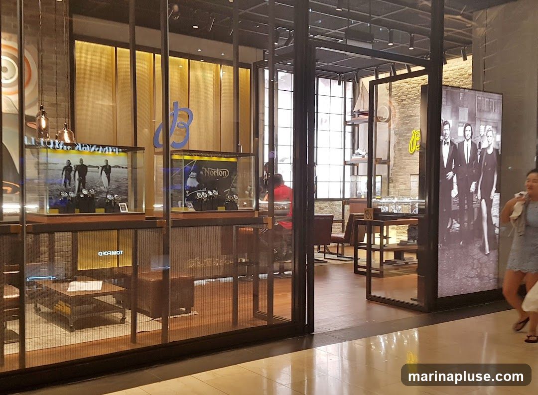 The interior of a modern watch store is visible, featuring glass display cases filled with watches. One case shows a promotional image of surfers, while another displays watches with a "Norton" logo. A large black and white poster of three people in formal wear stands near the entrance. The store has a warm, inviting ambiance with wooden floors and accents, and industrial-style lighting. A person in a red shirt is seen behind a counter.