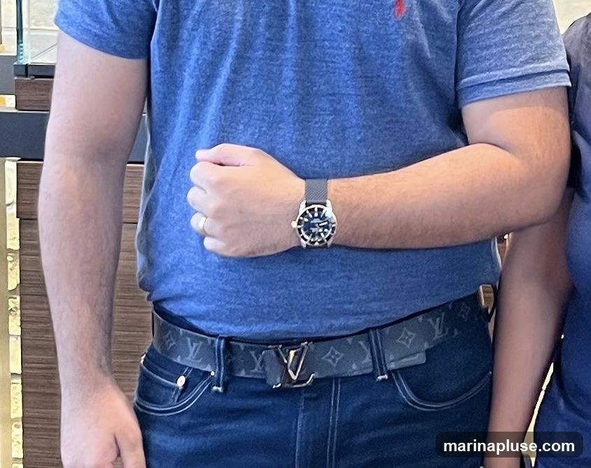 A man wearing a blue polo shirt, dark wash jeans, a watch on his left wrist, and a designer belt with a prominent logo. The background appears to be an indoor setting with wooden and metallic accents.