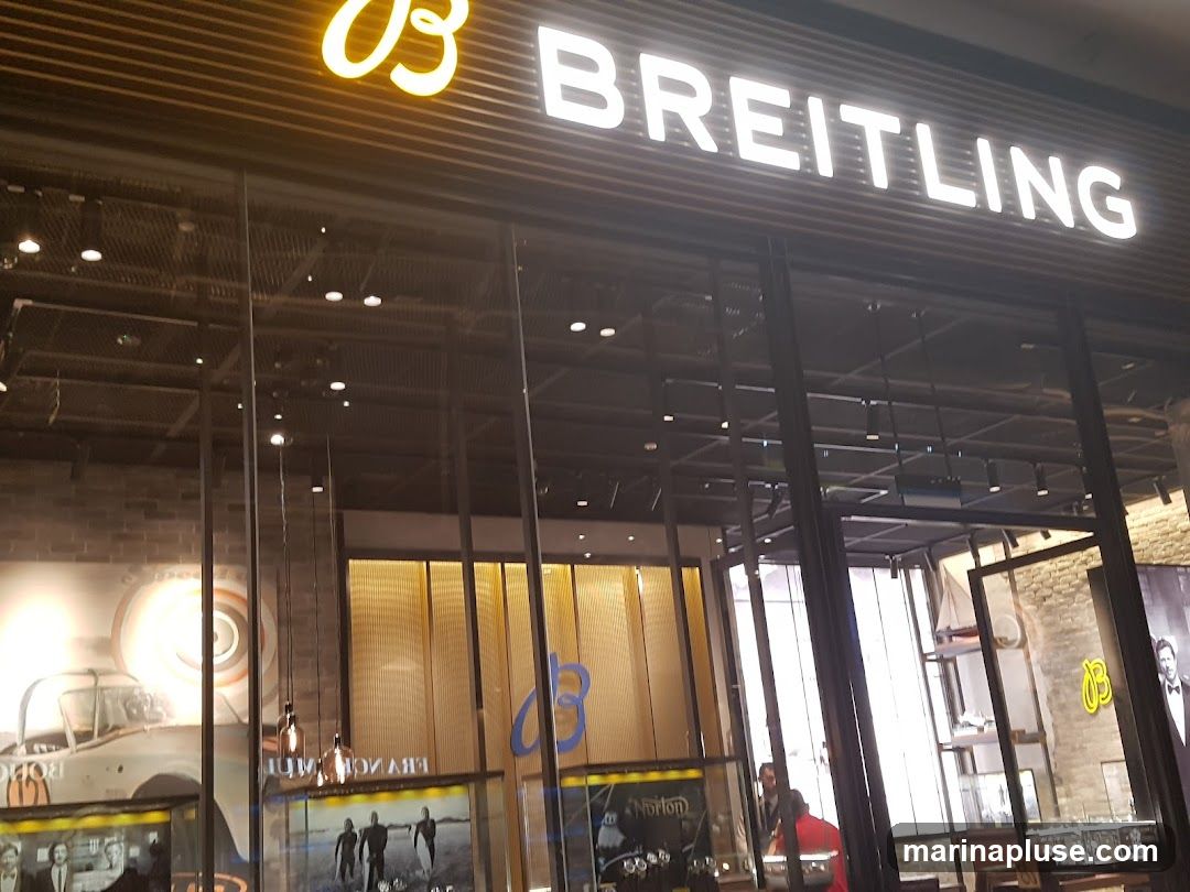 The exterior of a Breitling store with the brand name prominently displayed in illuminated white lettering above a glass facade. Inside, displays of watches and promotional imagery are visible, including a vintage car advertisement.