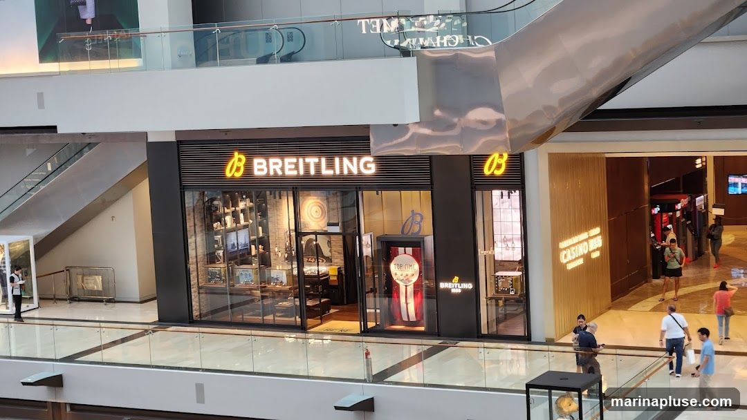 The exterior of a Breitling watch store is visible, with its prominent illuminated logo and name above the entrance. The store features large glass windows displaying watches and merchandise inside. A display case with a red and white striped background and the text "TOP TIME" is positioned near the entrance. Other shops, including a casino, are visible in the background.
