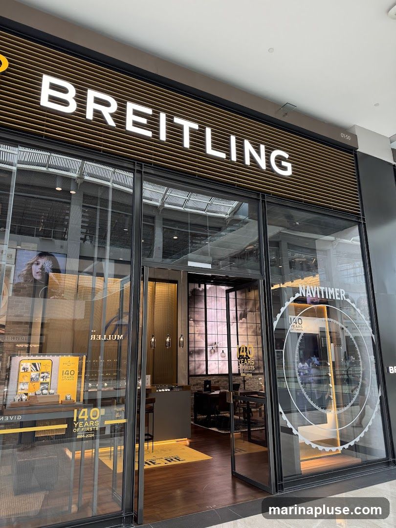 The exterior of a Breitling watch store. The large white "BREITLING" sign is visible above the entrance. The storefront features large glass windows displaying watches and promotional materials, including a "140 YEARS OF FIRSTS" advertisement. The entrance is open, revealing the interior of the store with display cases and seating areas.