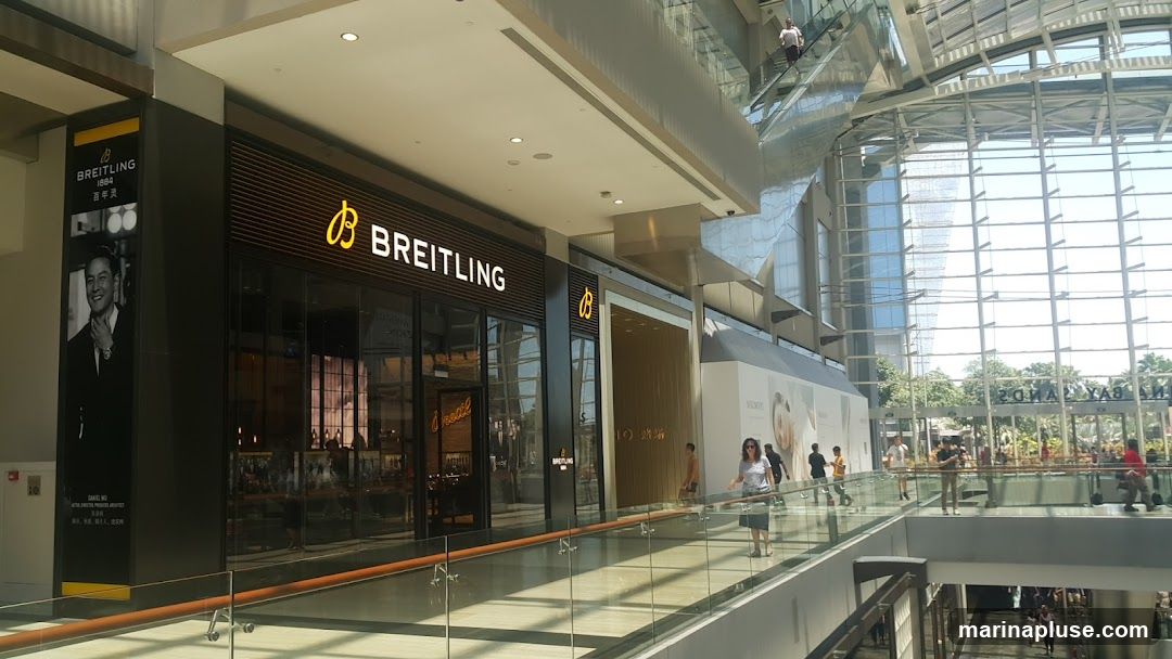 The exterior of a Breitling store in a modern shopping mall. The store has a large glass facade with the Breitling logo and name prominently displayed. To the left of the store entrance is a large vertical banner featuring a black and white image of a man wearing a watch, with text in English and Chinese. Inside the store, watches are displayed on shelves. The mall features a glass ceiling and walkways with railings.