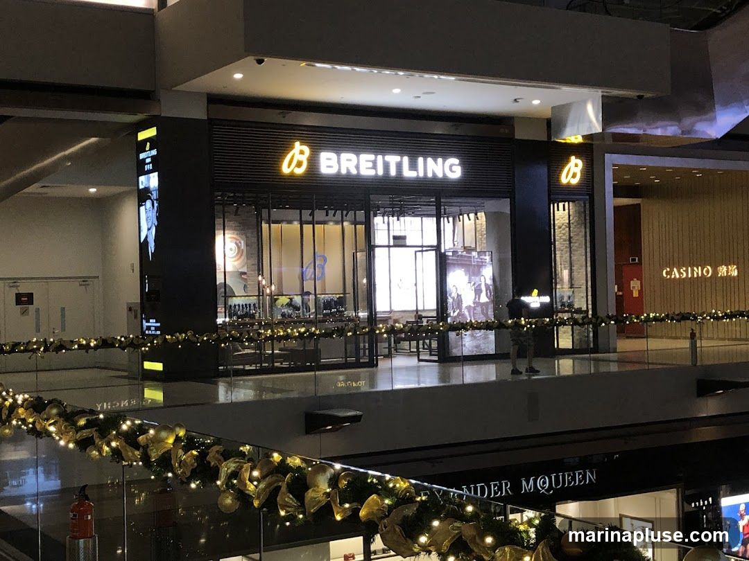 The exterior of a Breitling store in a modern shopping mall. The store has a dark facade with large glass windows displaying watches. The Breitling logo and name are prominently displayed in illuminated lettering above the entrance. To the right of the Breitling store is a casino entrance. Festive garlands with lights and gold ornaments are visible in the foreground, suggesting a holiday season.