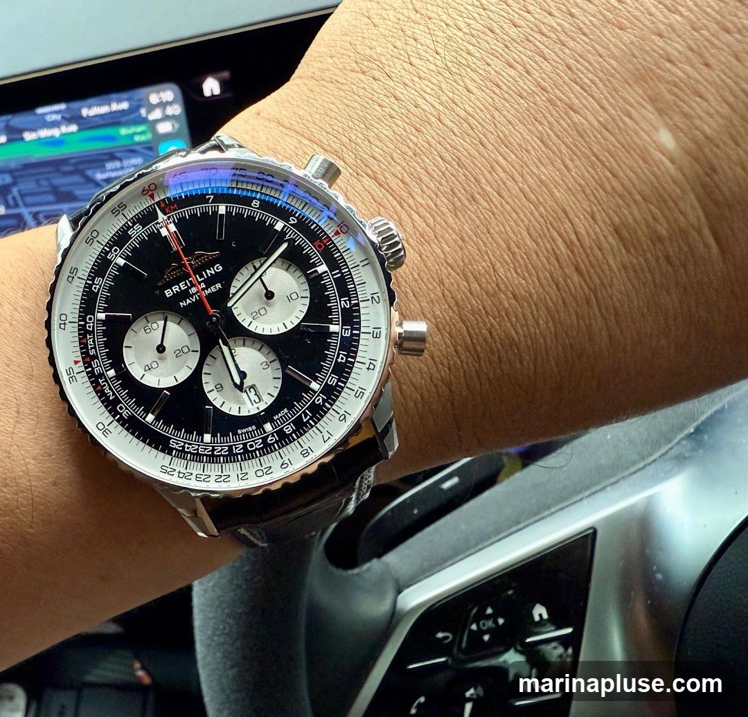 A close-up shot of a Breitling Navitimer watch on a person's wrist. The watch has a black dial with silver sub-dials and a silver bezel with black markings. The watch is worn in the context of being inside a car, with a steering wheel and a navigation screen visible in the background.