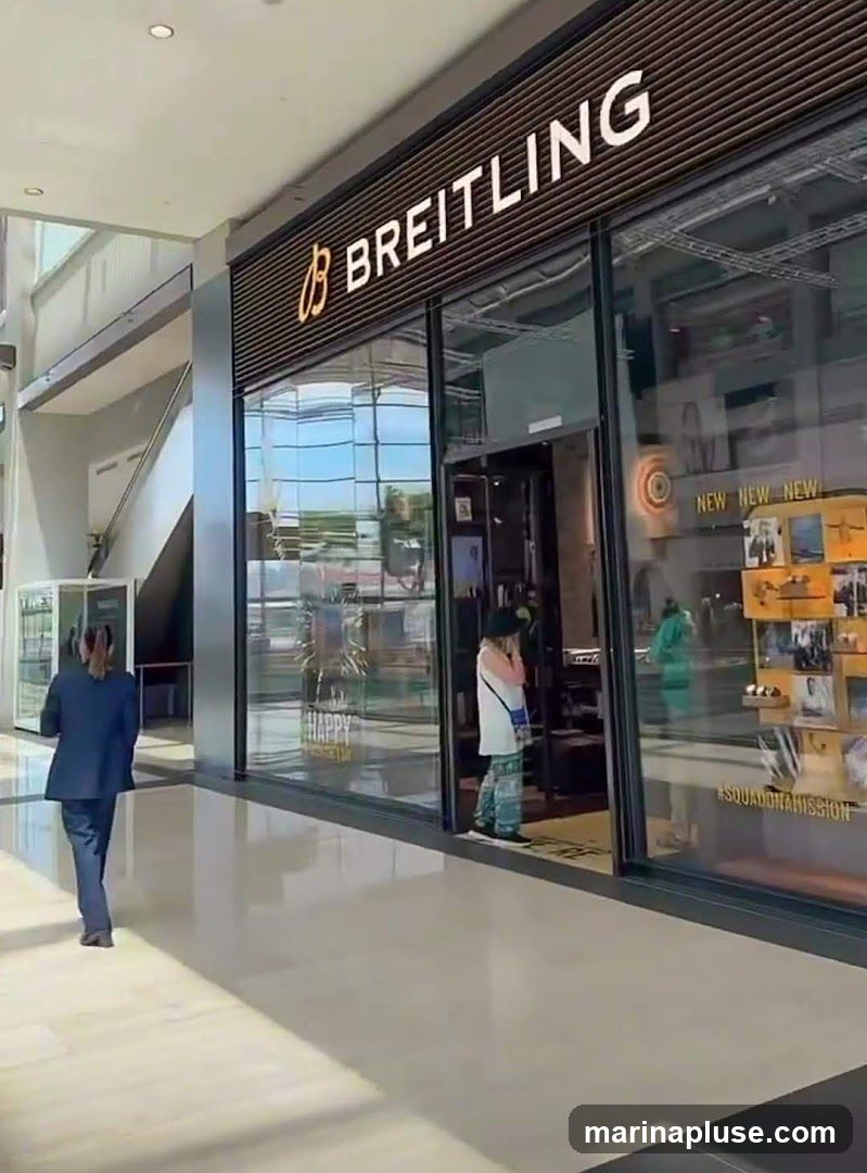 The exterior of a Breitling store in a mall. The store has a large glass facade with the Breitling logo and name prominently displayed above the entrance. Inside, displays of watches and promotional materials are visible. A person in a suit walks away from the store, and another person stands near the entrance.