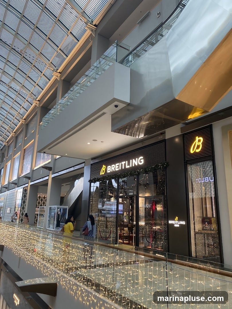 The exterior of a Breitling watch store in a modern shopping mall. The store has a large glass facade with the Breitling logo and name prominently displayed above the entrance. Inside, watches are displayed on shelves and in display cases. The storefront is decorated with festive string lights.