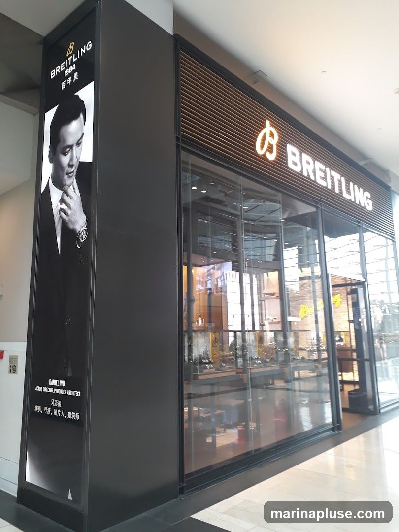 The exterior of a Breitling watch store. A large black pillar on the left features a black and white advertisement of Daniel Wu, with the Breitling logo and "1884 百年灵" text above him. The store itself has a large glass facade with the Breitling logo and name prominently displayed in illuminated white lettering above the entrance. Inside, display cases with watches are visible.