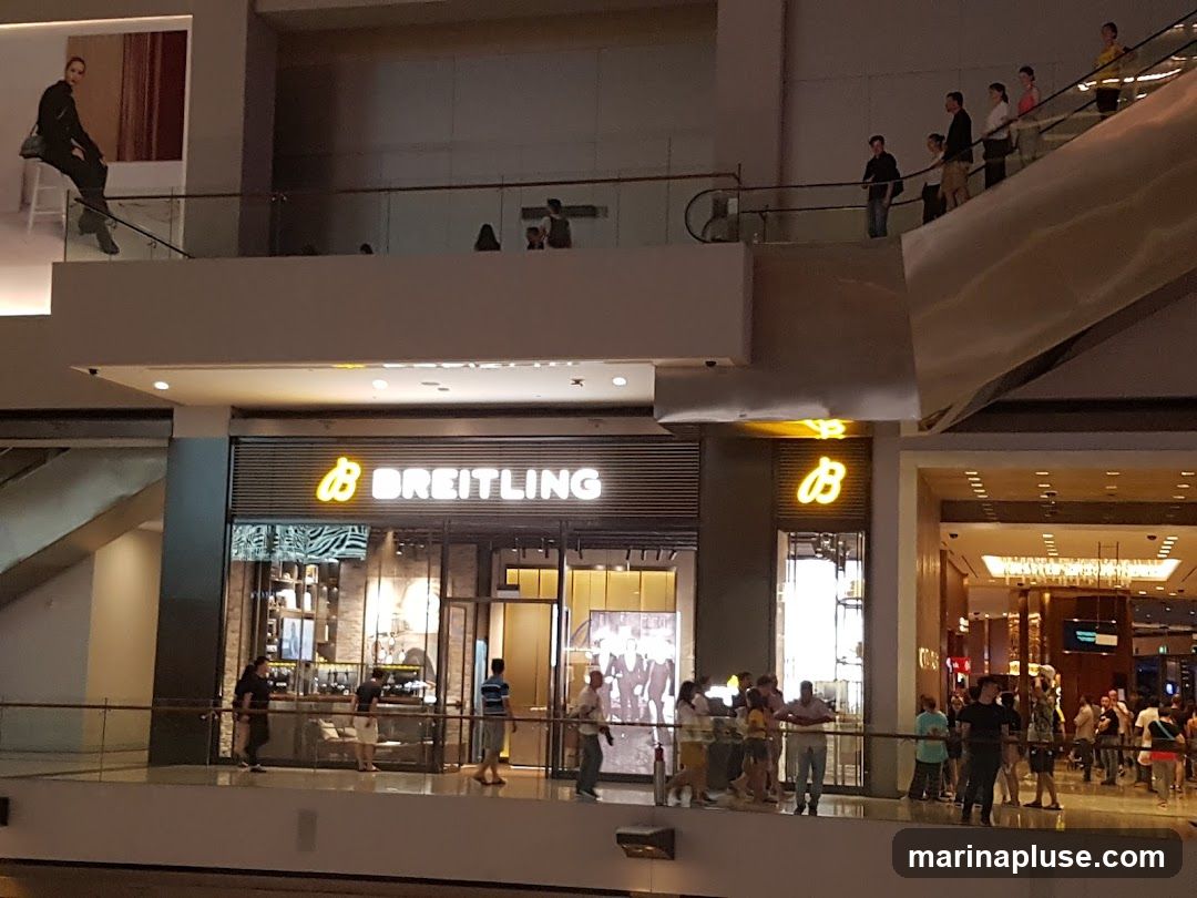 The exterior of a Breitling store is visible, with the brand name illuminated in yellow neon above the entrance. The store features a modern facade with large glass windows displaying watches and interior lighting. People are seen browsing outside the store and on escalators in the background.