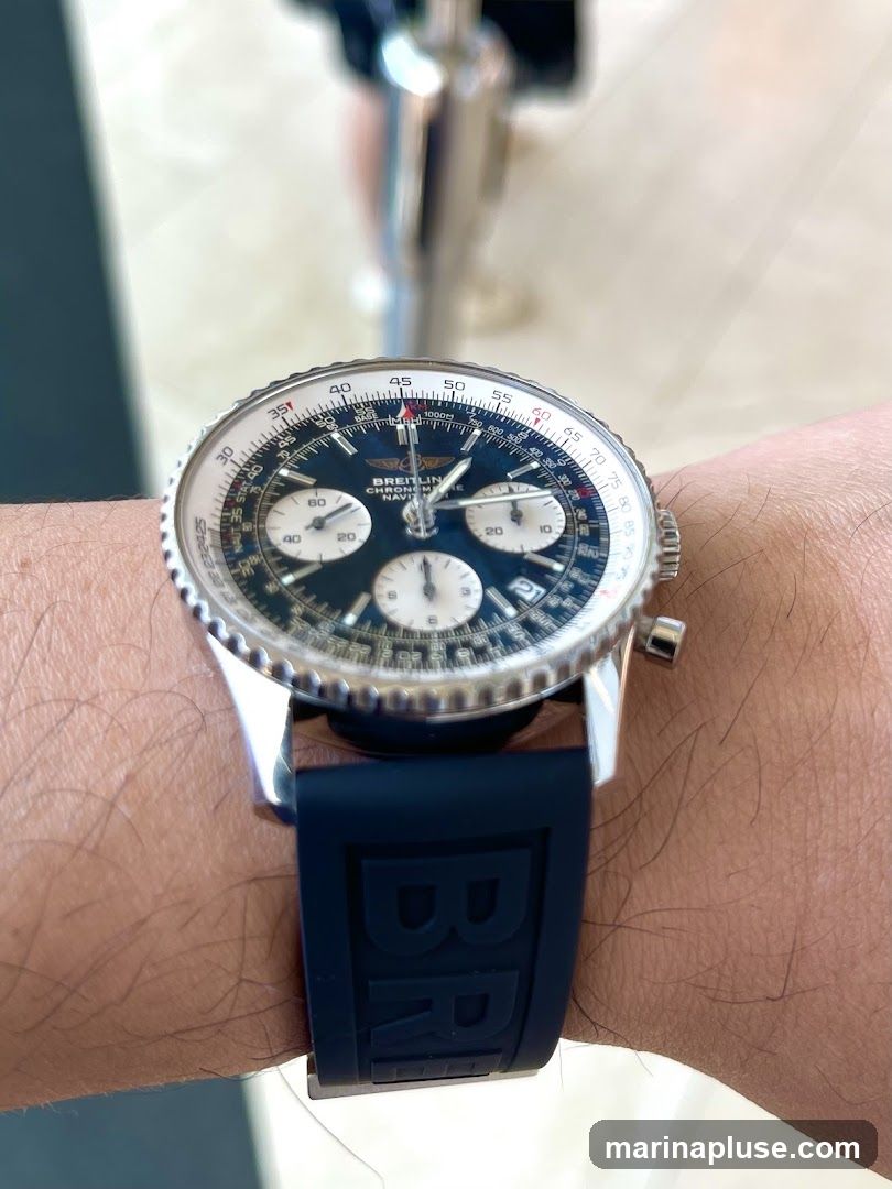 A close-up shot of a Breitling Chronomat Navitimer watch on a person's wrist. The watch has a dark blue dial with white sub-dials and a silver bezel. The strap is dark blue with embossed Breitling branding. The background is blurred, showing a metallic pole and a light-colored surface.