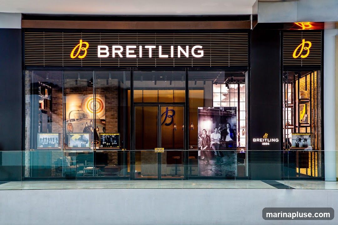 The exterior of a Breitling store with large glass windows and a prominent sign displaying the Breitling logo and name. Inside, displays of watches and artwork are visible. A large poster featuring three men is placed outside the entrance.