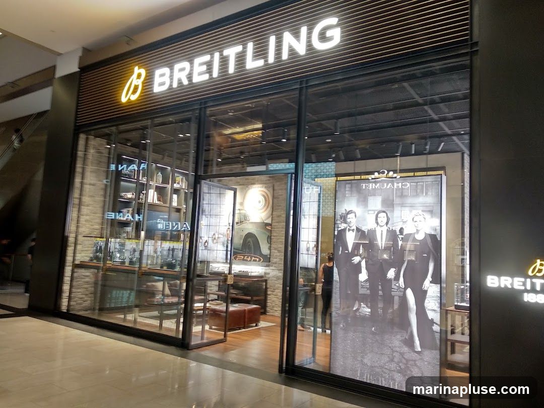 The exterior of a Breitling store with large glass windows and a prominent sign above. Inside, display shelves with watches and decorative items are visible. A large advertisement featuring three people in formal attire is displayed in one of the windows.